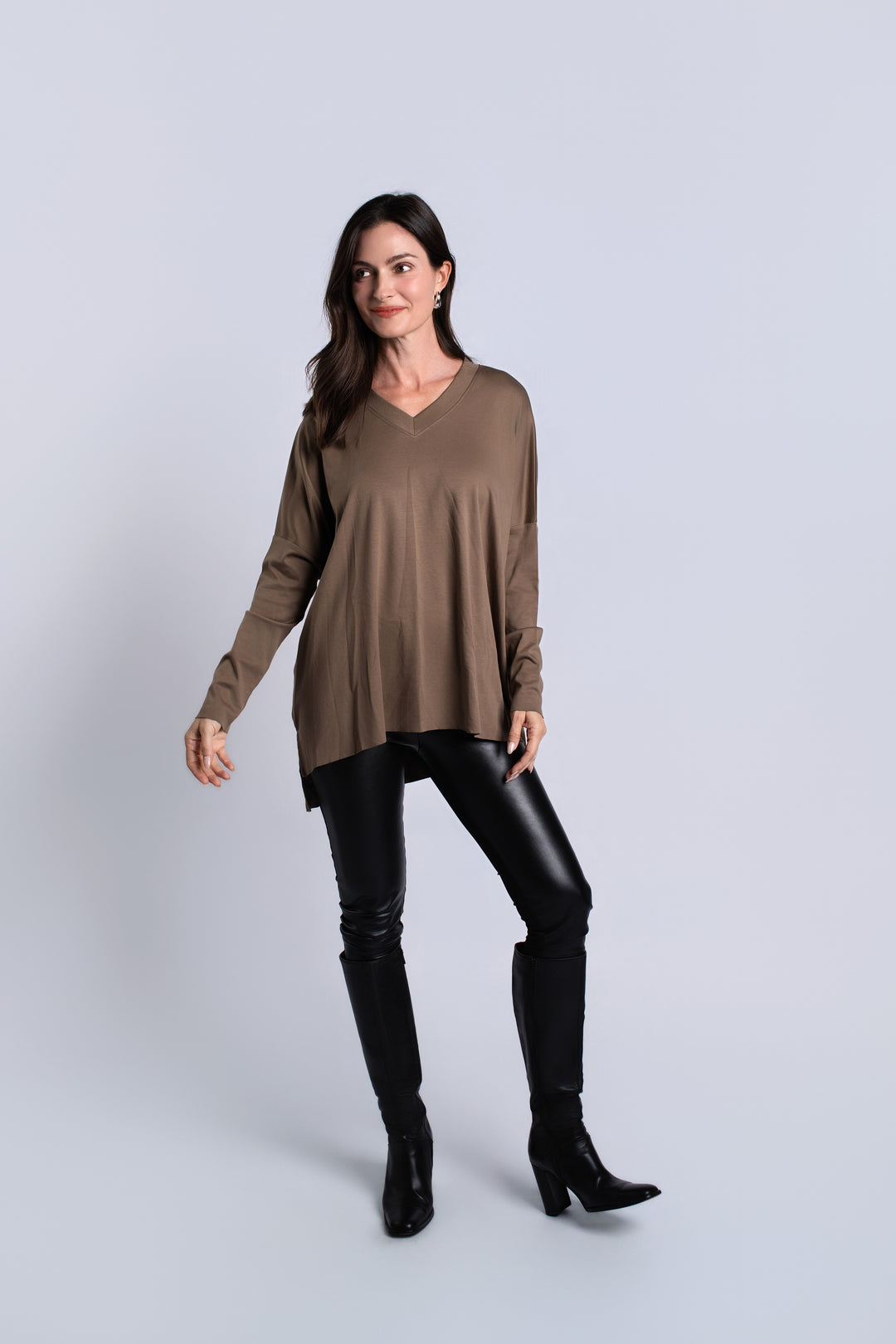 A woman modeling a taupe brown long-sleeve V-neck tunic top, paired with black faux-leather pants and black knee-high boots, against a light gray studio background.