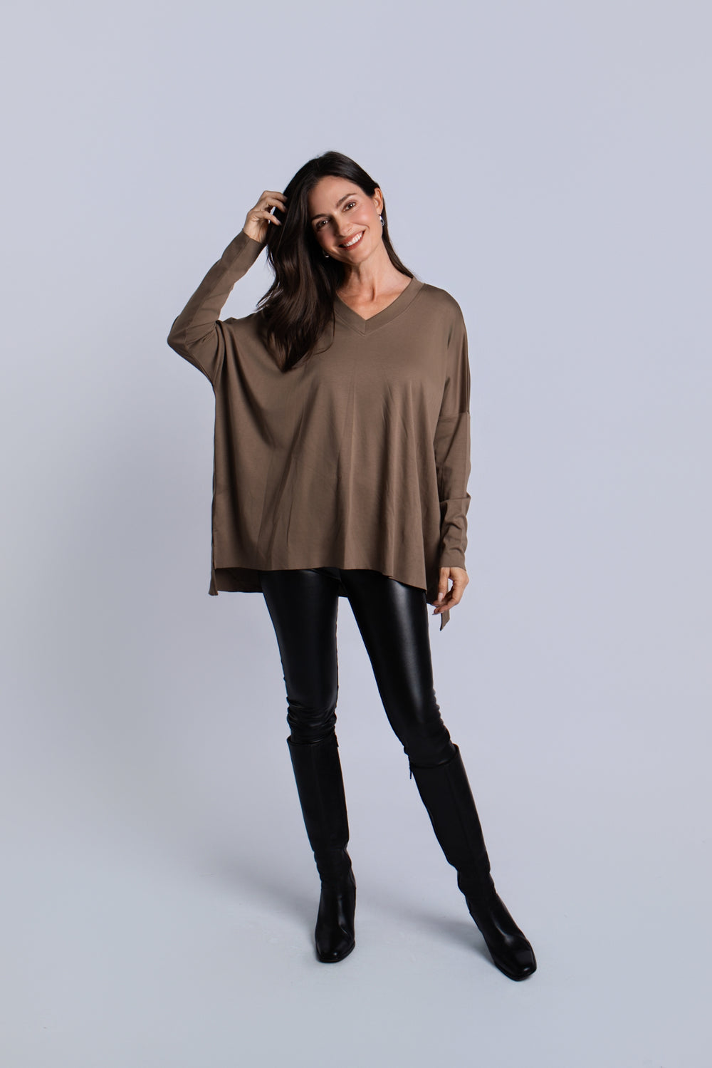 A woman modeling a loose, long-sleeve taupe-brown pima cotton V-neck top paired with black faux-leather pants and black knee-high boots, standing against a light gray studio background.