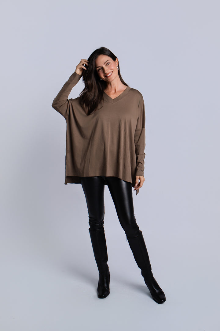 A woman modeling a loose, long-sleeve taupe-brown pima cotton V-neck top paired with black faux-leather pants and black knee-high boots, standing against a light gray studio background.