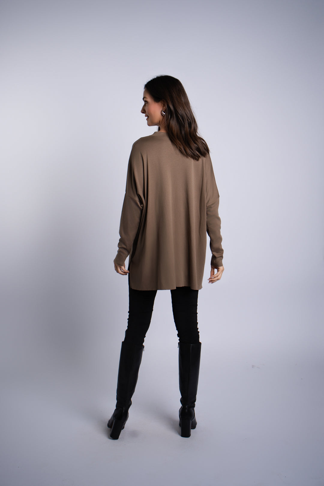 A woman is shown from the back wearing an oversized turtle-brown long-sleeve pima cotton top, black vegan leather leggings, and black knee-high boots against a light gray studio background.