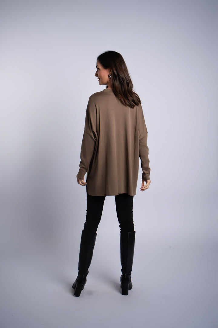 A woman is shown from the back wearing an oversized turtle-brown long-sleeve pima cotton top, black vegan leather leggings, and black knee-high boots against a light gray studio background.
