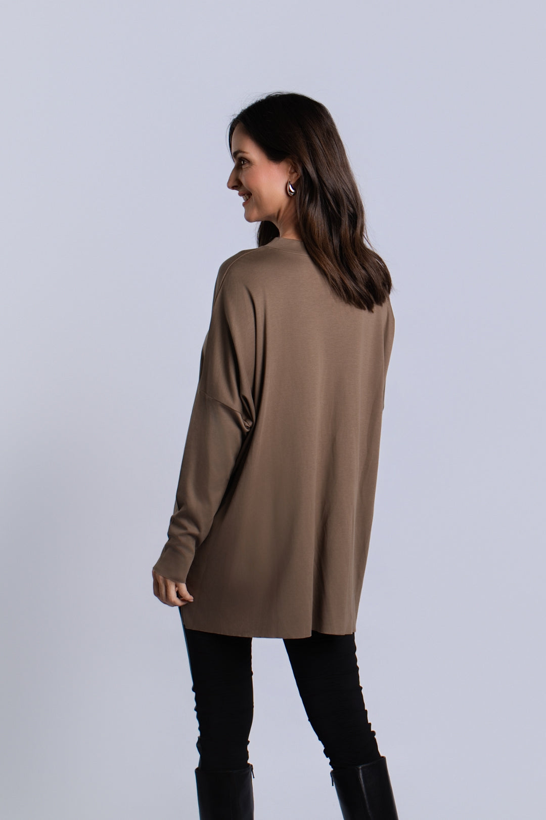 A woman wearing a turtle brown long-sleeve v-neck pima cotton top, black leggings, and black boots, shown from a side-back angle against a light gray studio background.