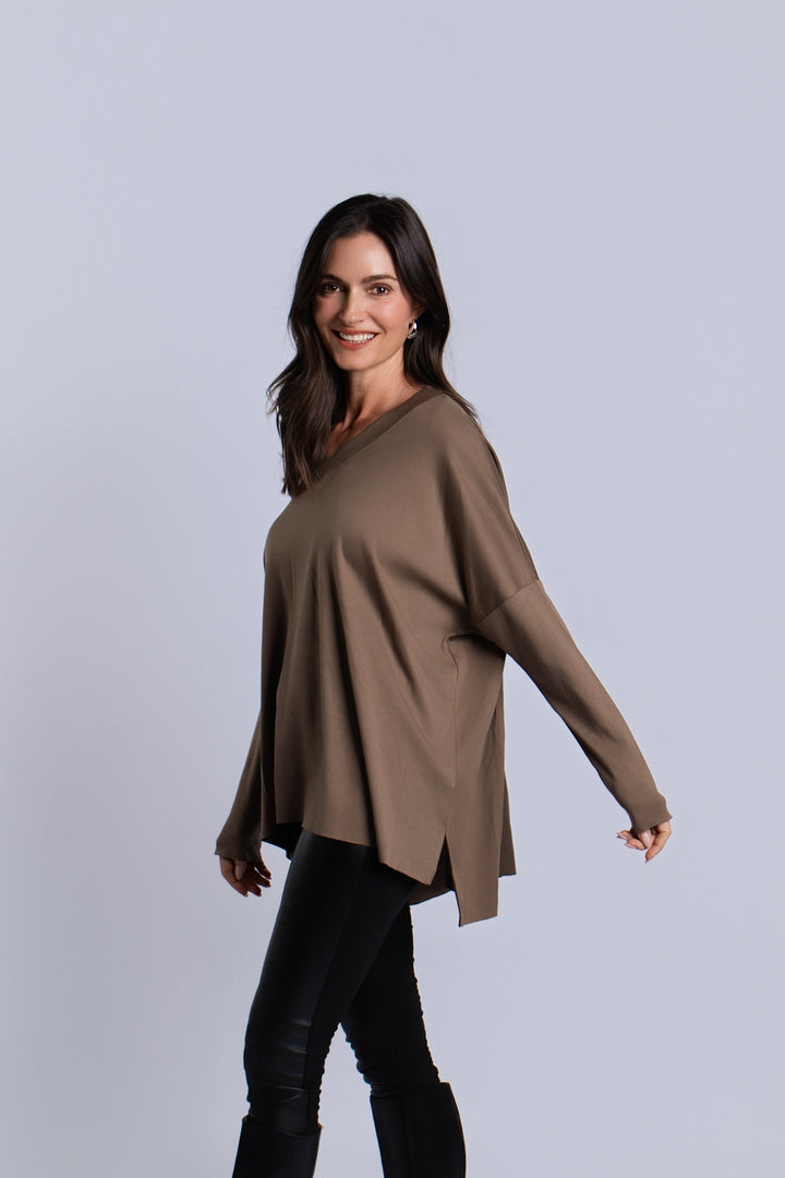 A woman modeling a loose, long-sleeve V-neck pima cotton top in taupe brown, paired with black pants, against a solid light gray studio background.